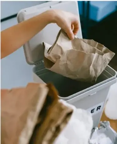 Mano depositando una bolsa de papel kraft en un contenedor de reciclaje metálico.