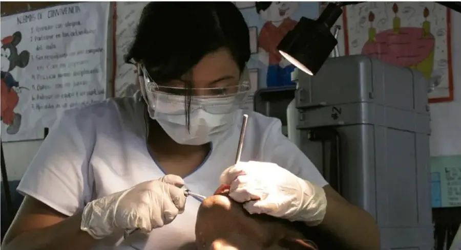 Fotografía de una odontóloga con mascarilla, careta y guantes realizando una intervención dental a un paciente, con equipo dental e iluminación profesional.