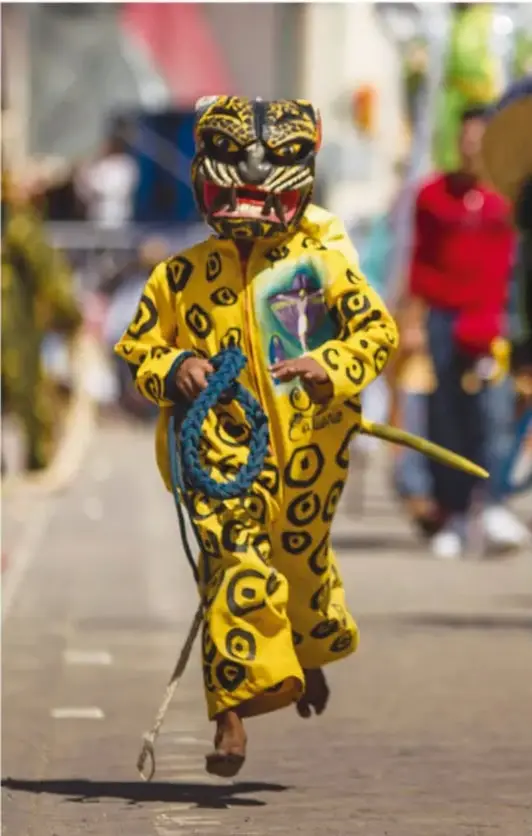 Danzante en movimiento con máscara de felino (jaguar o tigre) y traje amarillo con manchas negras, descalzo, corre sosteniendo una cuerda azul en una calle durante una festividad.