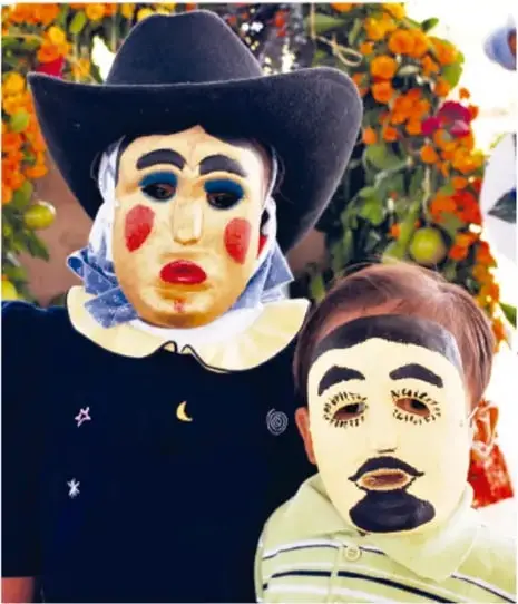 Persona adulta con sombrero negro y traje con estrellas, y un niño, ambos con máscaras pintadas de rostro claro, cejas marcadas, mejillas rojas y bigote; al fondo flores de cempasúchil. Foto: Jesús Eduardo Arteaga Flores, Tamaulipas.