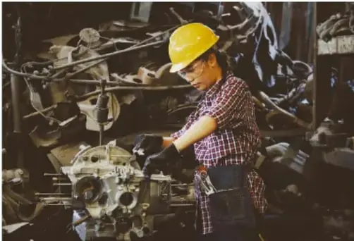 Fotografía de una trabajadora con casco amarillo operando maquinaria pesada en un taller industrial, ilustrando un sistema técnico en acción.
