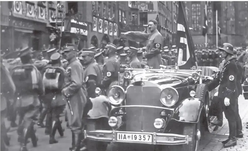 Fotografía en blanco y negro de un desfile nazi con vehículos y militares, con esvásticas en banderas y uniformes.