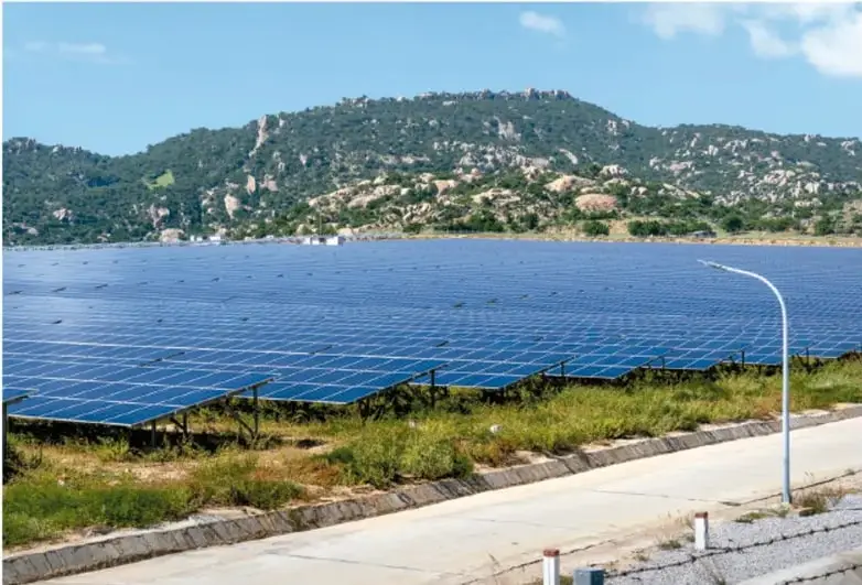 Fotografía de un parque solar fotovoltaico con paneles azules en primer plano, vegetación seca alrededor, una carretera y una lámpara curva; al fondo, montañas verdes bajo un cielo despejado.