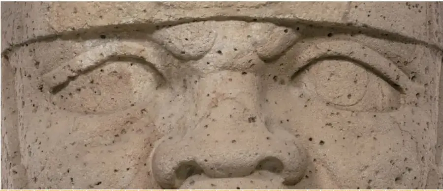 Fotografía de acercamiento a una cabeza colosal olmeca que muestra sus ojos, ceño fruncido y nariz tallados en la piedra volcánica llena de pequeños poros.