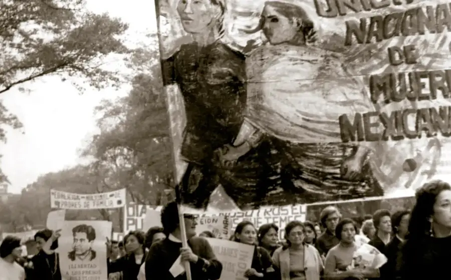 Fotografía en sepia de una manifestación multitudinaria con una gran pintura-banderola que muestra a dos mujeres y el texto 'Unión Nacional de Mujeres Mexicanas'; participantes portan carteles, entre ellos uno con el rostro de un hombre y la leyenda 'Libertad a los presos políticos'.