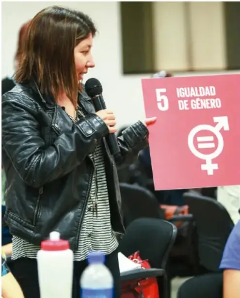 Fotografía de una mujer joven con chaqueta de piel sosteniendo un micrófono y un cartel rosa con el número 5 y la leyenda 'IGUALDAD DE GÉNERO' junto al símbolo de género con signo igual.