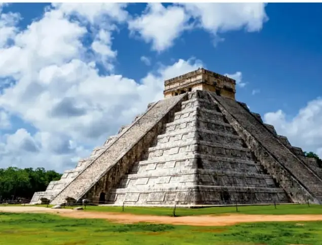 Fotografía de la pirámide de Kukulkán (El Castillo) en Chichén Itzá, Yucatán, con cielo azul y nubes, rodeada de árboles y pasto.
