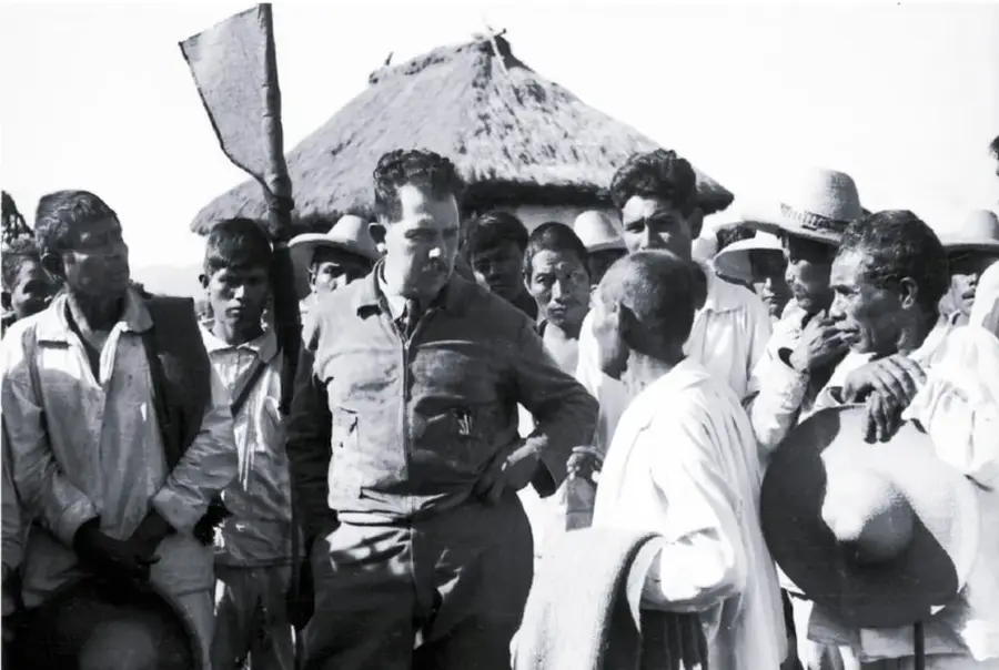 Fotografía en blanco y negro de Lázaro Cárdenas rodeado de campesinos, uno de ellos sostiene un sombrero; al fondo se observa una construcción con techo de palma.