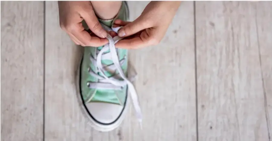 Fotografía en primer plano de las manos de un niño amarrando las cintas de su tenis verde claro, sobre un piso de madera.