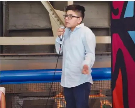 Fotografía de un estudiante presentando con micrófono frente a una audiencia, con pintura mural de fondo.