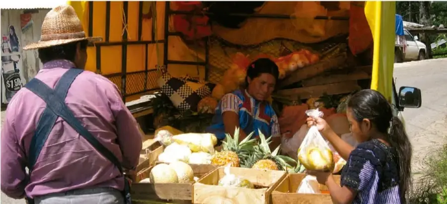 Fotografía de una venta comunitaria al aire libre: una mujer indígena entrega productos (piñas y frutas) a una niña mientras un hombre con sombrero de paja observa, con un puesto de mercado y un vehículo al fondo.