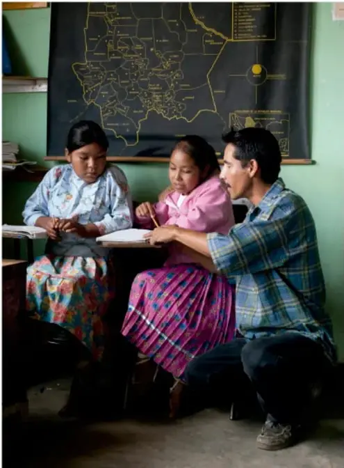 Fotografía de un docente y dos estudiantes indígenas sentados frente a un libro, con un mapa de México al fondo; crédito: Francisco Manuel Palma Lagunas, Líbano de México.
