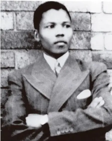 Fotografía en blanco y negro de Nelson Mandela joven, con traje y corbata, apoyado contra una pared de ladrillos.