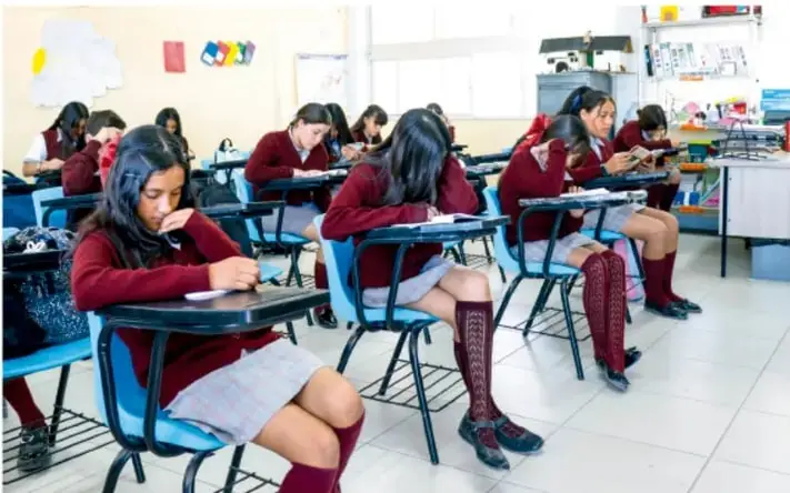 Fotografía de un aula de telesecundaria con estudiantes de uniforme (chamarra y falda/saco rojo vino, medias rojas) sentadas en pupitres individuales, trabajando de forma individual en sus cuadernos o libros.