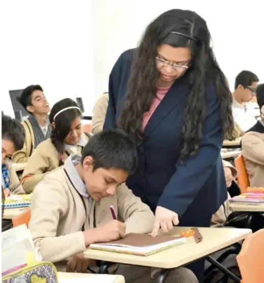 Fotografía de una docente de cabello largo ayudando a un estudiante a escribir en su cuaderno, mientras otros alumnos trabajan en equipos en el aula con computadoras.