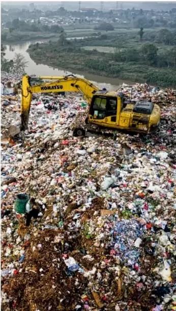 Fotografía de una excavadora amarilla trabajando en un tiradero de basura a cielo abierto, con un río contaminado al fondo.