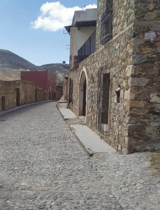 Calle empedrada de Real de Catorce junto a una casona de piedra con puerta en arco, rejas y balcón; cerros áridos al fondo.