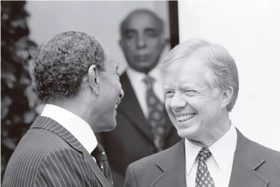 Fotografía en blanco y negro de dos hombres de traje sonriendo; uno de ellos es Jimmy Carter. Al fondo se observa otra persona.