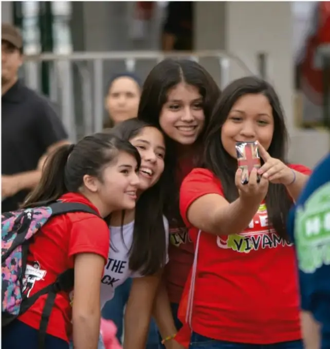 Fotografía de cuatro estudiantes sonrientes en grupo; una de ellas sostiene un teléfono celular para tomarse una selfie. Visten camisetas rojas y cargan mochilas, en un espacio exterior con personas al fondo.