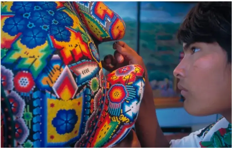 Fotografía en color de un joven huichol observando una pieza de arte tradicional de chaquira multicolor, con apaisado y marco blanco.