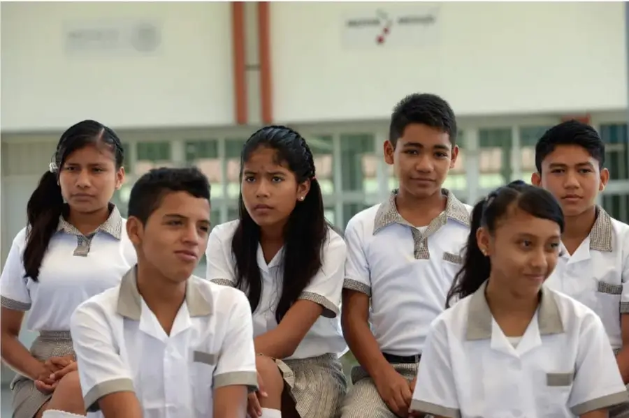 Fotografía de un grupo de seis adolescentes con uniforme escolar blanco, sentados en un patio con ventanales al fondo.