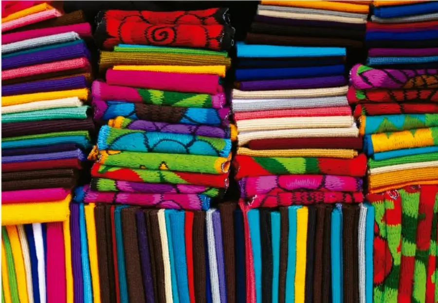 Fotografía de textiles y prendas tradicionales de colores vivos (rosa, rojo, azul, verde, amarillo, morado) dobladas y apiladas en columnas, con estampados y franjas, propias de artesanías textiles regionales de México.