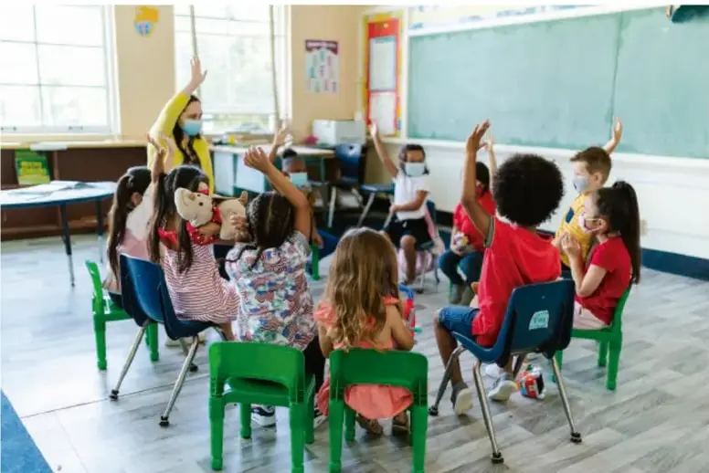 Fotografía de un grupo de estudiantes de primaria sentados en círculo en el aula, con una maestra de pie; varios niños levantan la mano para participar. Al fondo hay un pizarrón verde y ventanas.