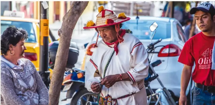 Fotografía callejera: un hombre mayor con traje tradicional mexicano (sombrero adornado con flores rojas, camisa blanca con bordado rojo y morral) conversa con una mujer sentada; a la derecha un joven con gorra y playera roja; al fondo autos, motocicletas y un taxista. Escena de convivencia cotidiana y diversidad cultural.