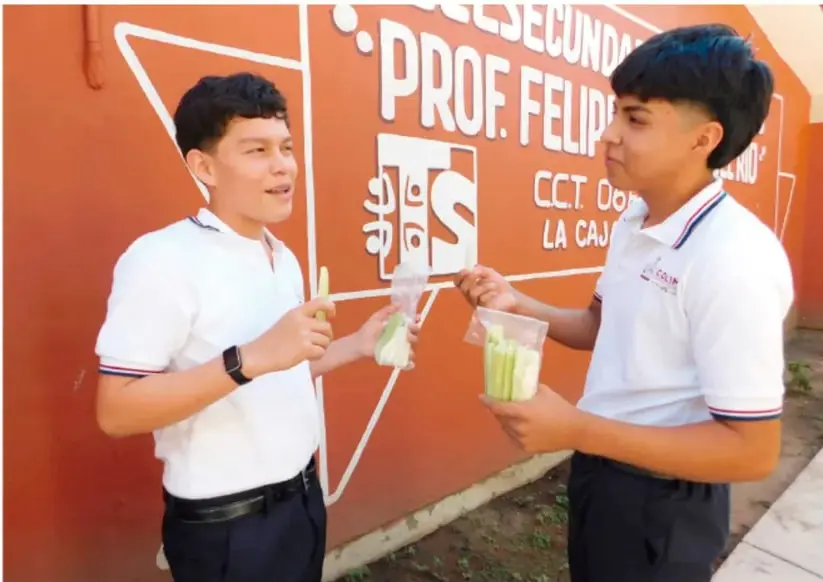 Fotografía de dos estudiantes de Telesecundaria con uniforme blanco, de pie frente a un muro naranja pintado con el nombre de su escuela ("...SECUNDA... PROF. FELIPE...") y logotipos; uno sostiene un recuerdo o producto del proyecto y el otro sostiene bolsas con objetos, en actitud de conversación.