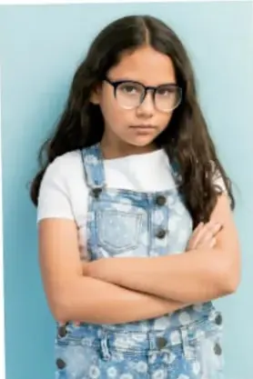 Fotografía de una niña con lentes y overol de mezclilla, con los brazos cruzados.