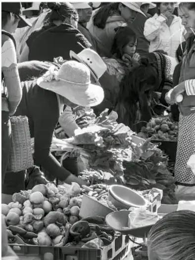 Fotografía en blanco y negro de un mercado o tianguis comunitario: varias personas, algunas con sombrero, revisan y venden productos como frutas, verduras y canastas colocadas en el suelo y en mesas.