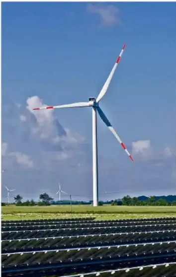 Fotografía de un campo con paneles solares en primer plano y aerogeneradores eólicos al fondo bajo cielo azul con nubes.