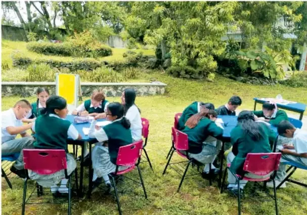 Fotografía de estudiantes de telesecundaria trabajando en grupos al aire libre, sentados en mesas rojas y azules en un área verde con árboles.