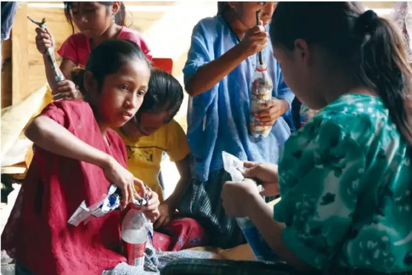Fotografía de niñas y niños trabajando en equipo con botellas de plástico y materiales para elaborar ecoladrillos en un espacio comunitario.