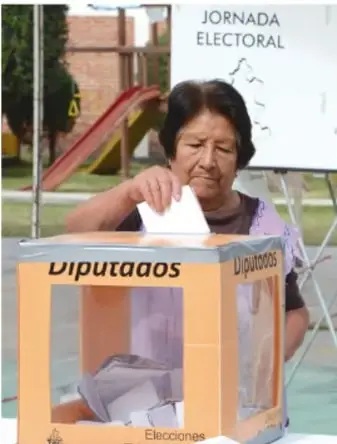 Mujer adulta deposita su voto en una urna transparente marcada 'Diputaos'; al fondo se lee 'JORNADA ELECTORAL'.