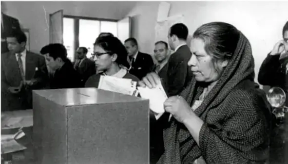Fotografía en blanco y negro de una mujer depositando su voto en una urna, acompañada de otras personas en una casilla electoral.