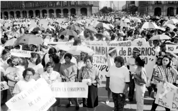 Fotografía en blanco y negro de una multitudinaria marcha ciudadana en una plaza; las personas portan pancartas con mensajes contra la corrupción y en defensa de derechos, bajo paraguas y sombrillas.