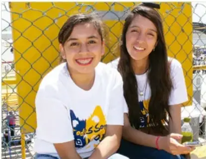 Fotografía de dos jóvenes sonrientes junto a una reja metálica amarilla, con camisetas blancas, ilustrando la integración y el bienestar en la comunidad.