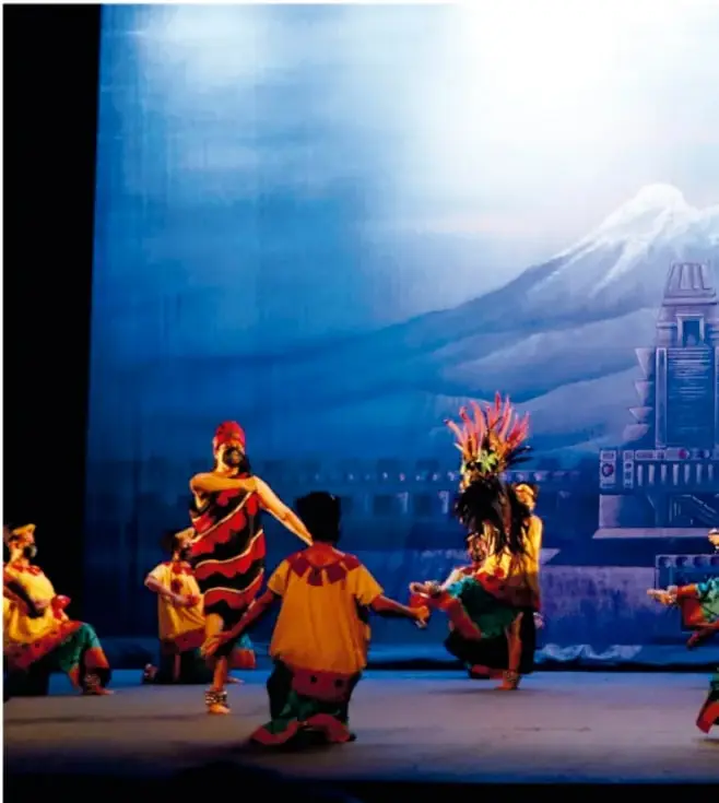 Fotografía de una danza escénica con bailarines de vestuarios coloridos (naranja, rojo, verde), algunos con penachos, sobre un telón de fondo azul con volcanes y pirámides pintados.