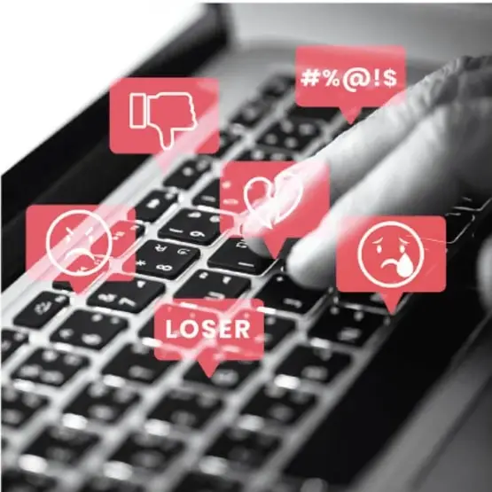 Fotografía de un teclado de computadora con íconos flotantes de agresión digital: pulgar abajo, símbolo de prohibido, corazón roto, carita triste, la palabra LOSER y el símbolo #%@!$.