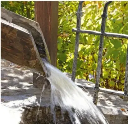 Fotografía de un chorro de agua cayendo desde una canaleta de madera hacia una superficie de piedra, con vegetación verde al fondo.