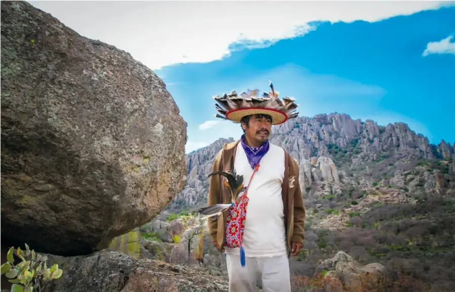 Fotografía de un mara'akame wixaritari en la sierra: lleva sombrero tradicional adornado con plumas y diseños, paliacate morado al cuello, morral bordado y plumas; junto a una gran roca, con montañas y cielo azul de fondo.