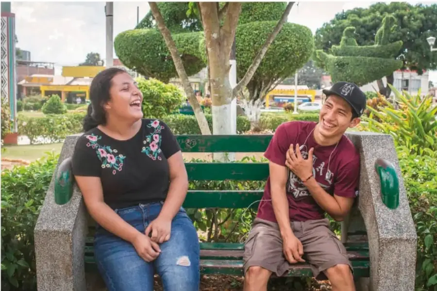 Chica y chico adolescentes riendo juntos sentados en una banca verde de un parque con jardines podados y edificios al fondo, mostrando una amistad respetuosa.