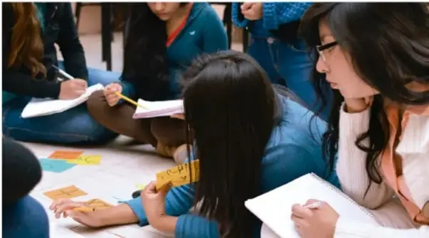 Fotografía de estudiantes sentadas en el piso trabajando con papeles de colores y cuadernos, colaborando en equipo.