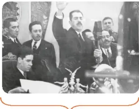 Fotografía en blanco y negro de la toma de protesta de Lázaro Cárdenas, con la banda presidencial y el brazo en alto.