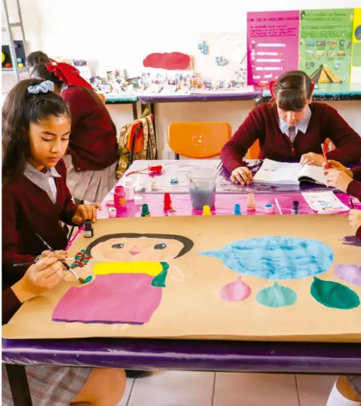 Fotografía de estudiantes de telesecundaria con uniforme bordó pintando con pinceles y temperas sobre una cartulina con figuras de colores y una charca azul, en un aula con materiales artísticos.