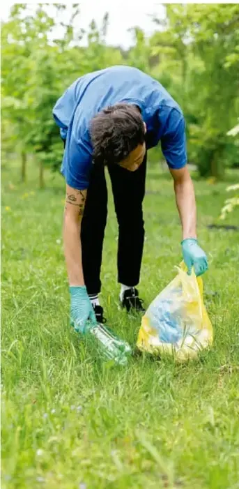 Fotografía de una persona joven con guantes recogiendo basura y plásticos del pasto, con una bolsa amarilla.