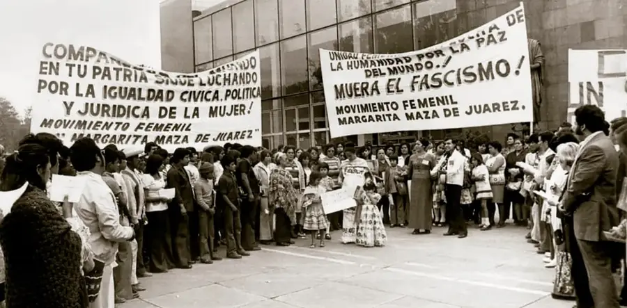 Fotografía en blanco y negro de una multitud en una explanada urbana con dos grandes carteles: uno dice 'COMPARTIENDO EN TU PATRIA SINCE LUCHANDO POR LA IGUALDAD CÍVICA, POLÍTICA Y JURÍDICA DE LA MUJER — MOVIMIENTO FEMENIL MARGARITA MAZA DE JUÁREZ' y el otro 'UNIDAD DE ... POR LA PAZ DE LA HUMANIDAD DEL MUNDO — ¡MUERA EL FASCISMO! — MOVIMIENTO FEMENIL MARGARITA MAZA DE JUÁREZ'. Personas de diversas edades observan la manifestación.