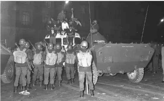 Fotografía en blanco y negro de soldados con equipo antidisturbios frente a un vehículo blindado durante una protesta.