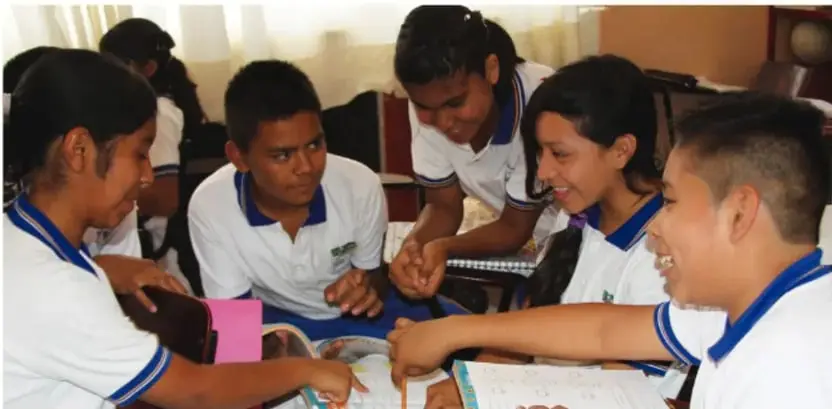 Fotografía de cinco estudiantes de secundaria con uniforme blanco y cuello azul, reunidos alrededor de una mesa, trabajando con libros y materiales en una actividad colaborativa.